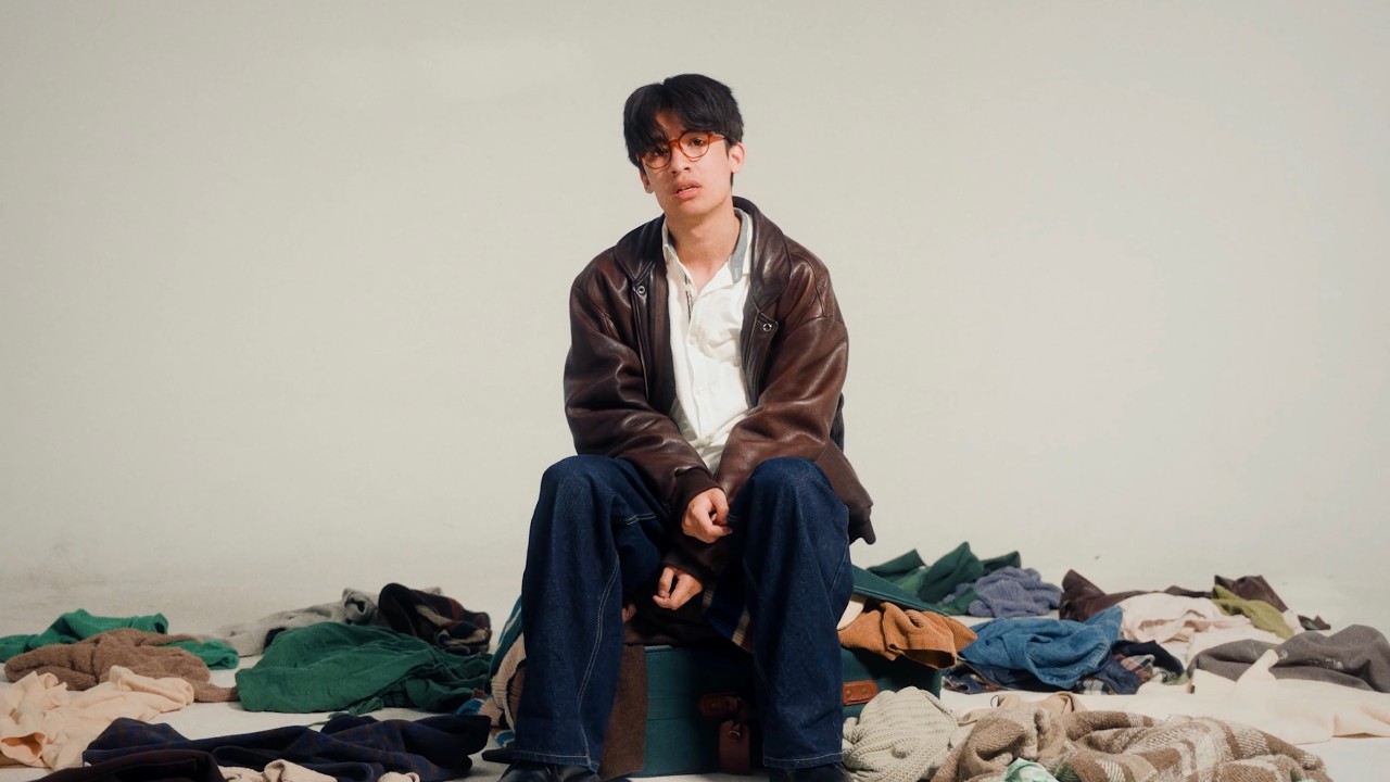 Still do videoclipe de HOMESICK: mala aberta e roupa espalhada no chão do estúdio